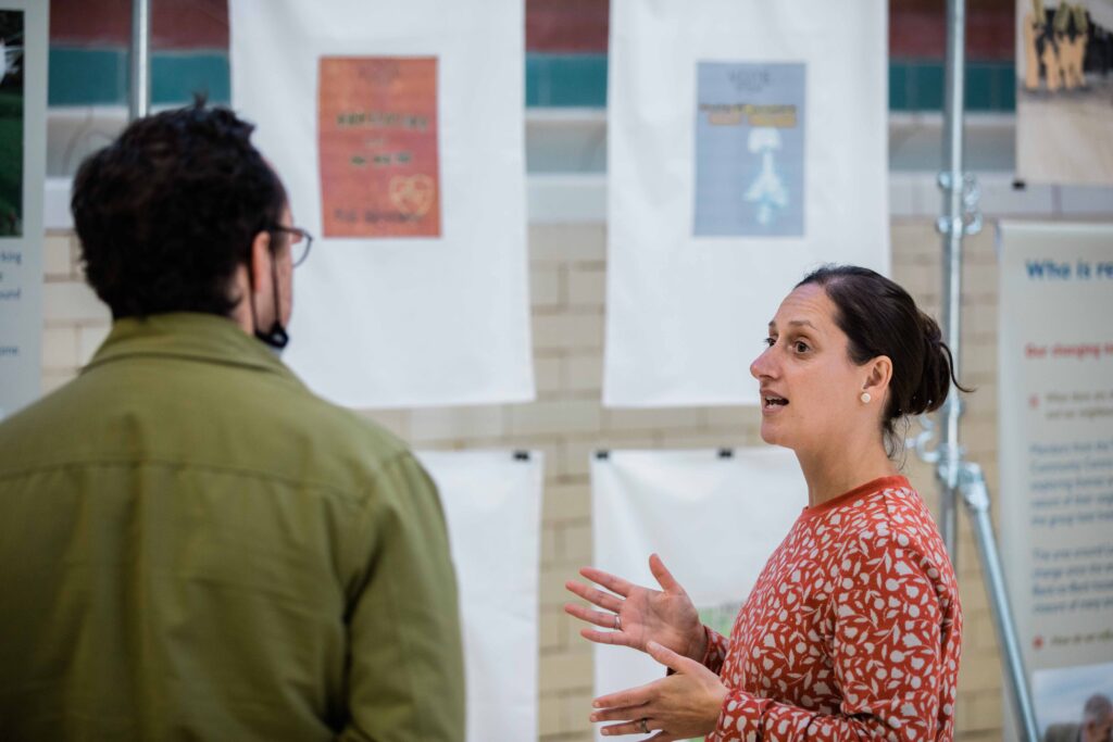 A white woman in her 30s speaks with a white man in his 30s in front of the exhibition textile banners in the deep end of an empty Edwardian swimming baths.